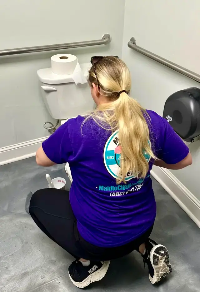 A cleaner crouched scrubbing a commercial restroom, the Maid to Clean logo on her shirt